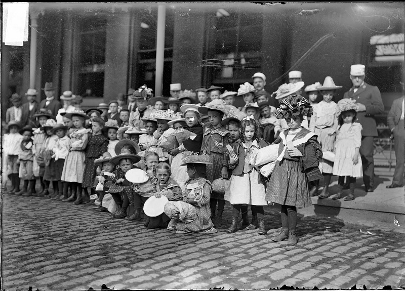 Girl wearing a ruffled hat in a group of children assembled for a fresh air outing. Image of many children, some with packages and bags, standing and sitting on a sidewalk and in a street, as part of a Chicago Daily News Fresh-Air Fund outing, possibly outside a train station in Chicago, Illinois. A few adults are standing with the children. Source: DN-0000076, Chicago Daily News negatives collection, Chicago History Museum. Date: ca. 1902.