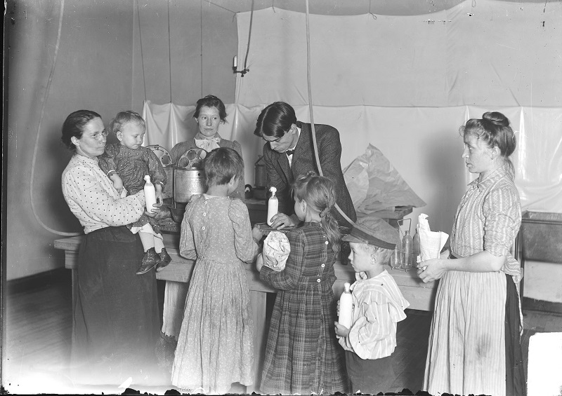 Sterilizing milk for children at Northwestern University settlement house. Image of women and children, some holding bottles of milk, standing around a table and a man standing behind the table at a Northwestern University settlement house in Chicago, Illinois. Source: DN-0000806, Chicago Daily News negatives collection, Chicago History Museum. Date: 1903 July 6.