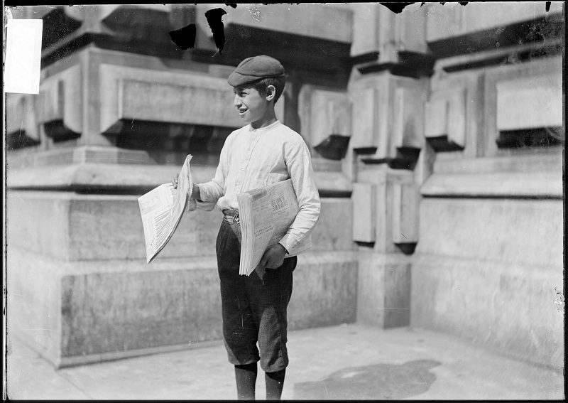 Newsboy selling newspapers. Image of a newsboy, with a stack of papers under his arm, holding a paper out on a commercial street in Chicago, Illinois. A pedestrian is visible in the foreground. Source: DN-0001792, Chicago Daily News negatives collection, Chicago History Museum. Date: 1904 Aug. 11.
