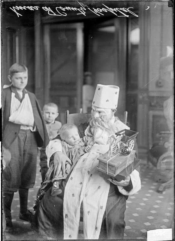 Christmas scene at Cook County Hospital, person dressed as Santa Claus holding a child on his lap with two other children standing nearby. Image of a Christmas scene at Cook County Hospital in Chicago, Illinois. A person dressed as Santa Claus holding a child on his lap with two other children standing nearby. Source: DN-0007024, Chicago Daily News negatives collection, Chicago History Museum. Date: 1909.