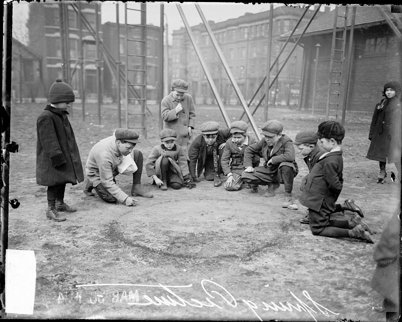 Seven boys kneeling, playing marbles on a playground, while two boys stand and look on, a girl is visible in the right on the image, swingsets are visible in the background. Group portrait of seven boys kneeling, playing marbles on a playground in Chicago, Illinois. Two boys stand and look on, a girl is visible in the right on the image, swingsets are visible in the background. Text on negative reads: Spring Picture. Source: DN-0062448, Chicago Daily News negatives collection, Chicago History Museum. Date: ca. 1914 Mar. 30.