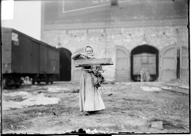 Woman carrying planks of wood outside a railroad depot. Image of one woman carrying wooden planks in a lot outside a railroad depot in Chicago, Illinois. She is walking toward the camera, away from a railroad depot. A railroad car standing outside the depot, and several trains inside the depot are visible in the background. Source: DN-0000502, Chicago Daily News negatives collection, Chicago History Museum. Date: ca. 1903.