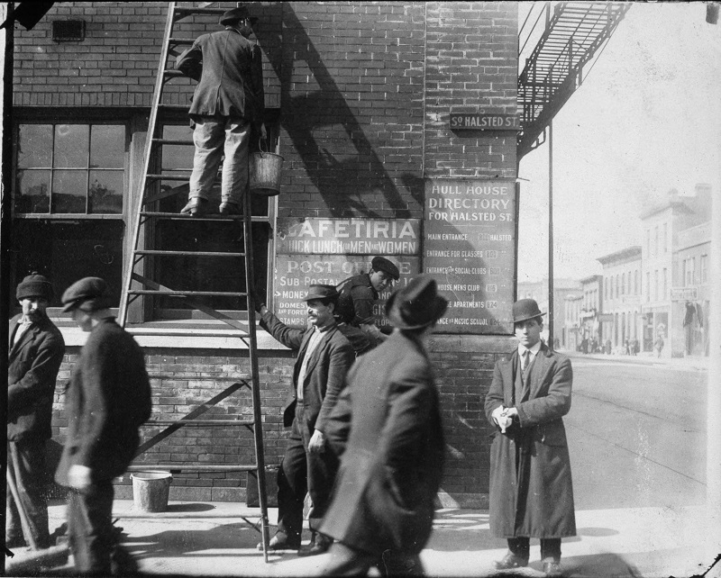 Corner view of Hull House building showing Hull House directory, cafeteria signs, men working and standing about. Description: Hull House building showing Hull House directory, cafeteria signs, men working and standing about; Chicago, IL. Source: ICHi-01542. Chicago History Museum. Reproduction of photographic print, photographer unknown. Date: n.d.
