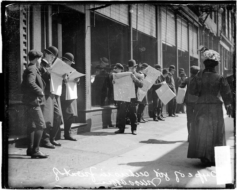Job seekers, men standing in front of the Chicago Daily News building, looking at newspapers with pedestrians walking nearby. Image of job seekers, men standing in front of the Chicago Daily News building, looking at newspapers with pedestrians walking nearby. The building was located at 15 North Wells Street (formerly 123 Fifth Avenue) in the Loop community area of Chicago, Illinois. Source: DN-0051332, Chicago Daily News negatives collection, Chicago History Museum. Date: 1907.